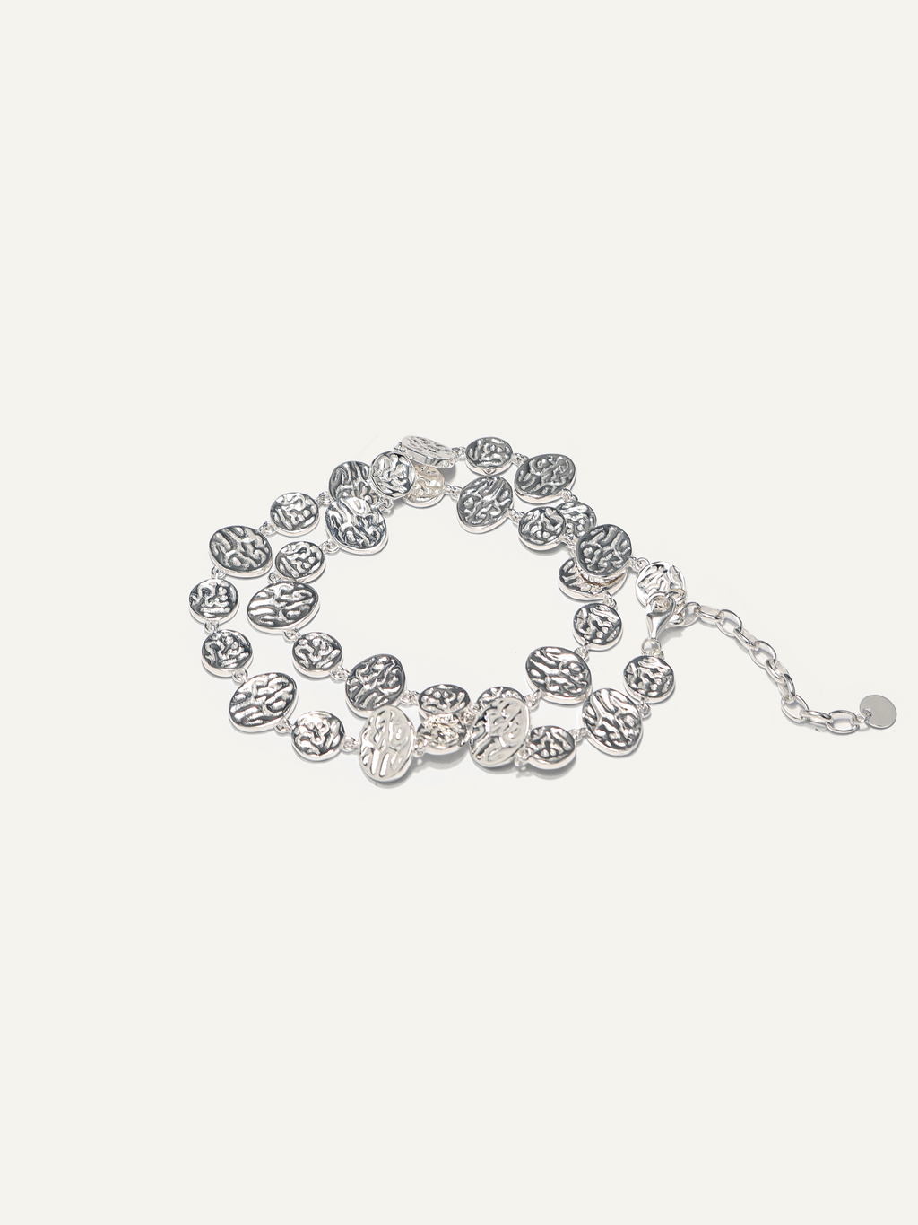A silver-toned multi-strand bracelet featuring textured, coin-like oval links and a small chain extender, displayed on a light cream background.