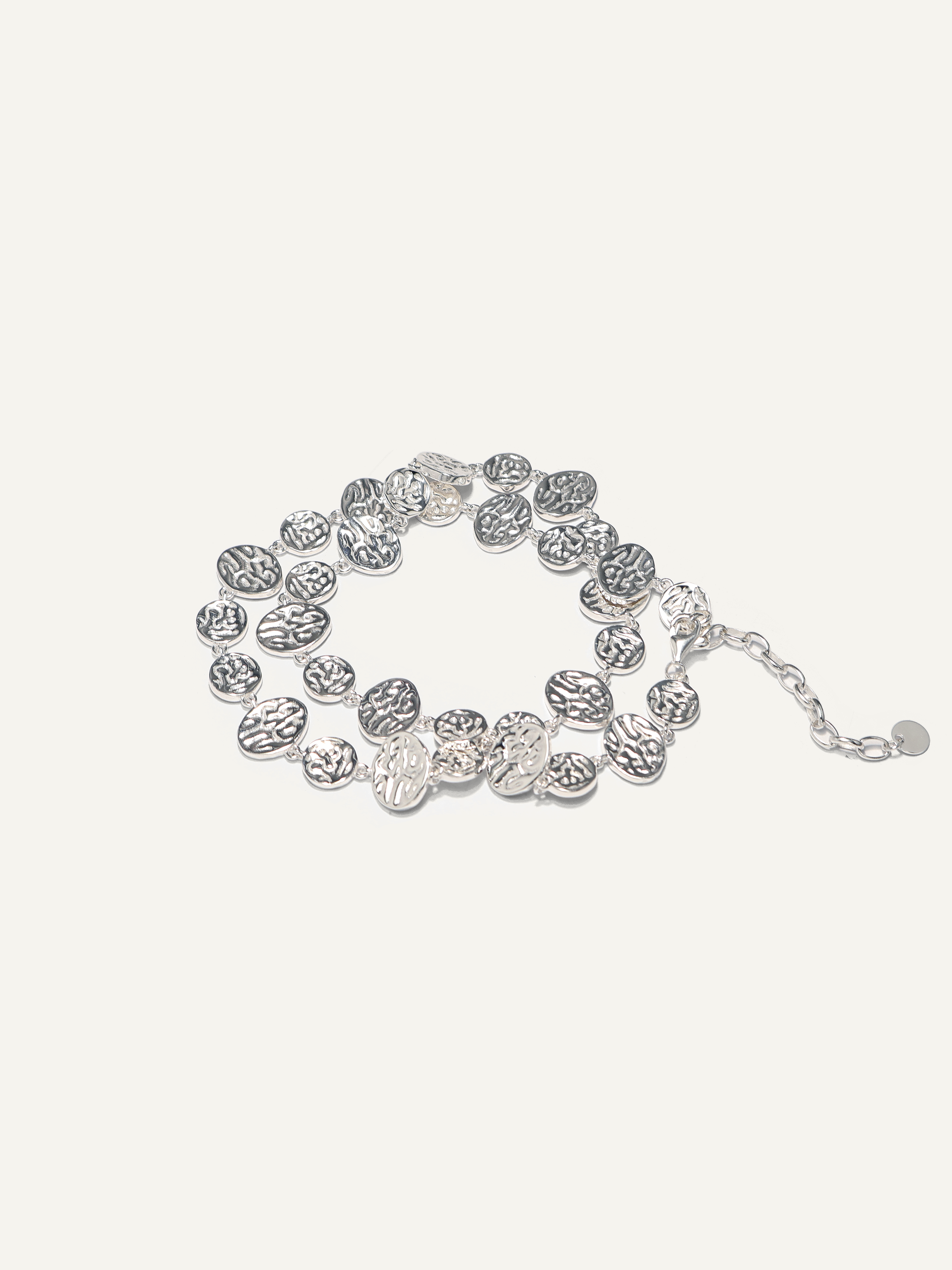 A silver-toned multi-strand bracelet featuring textured, coin-like oval links and a small chain extender, displayed on a light cream background.