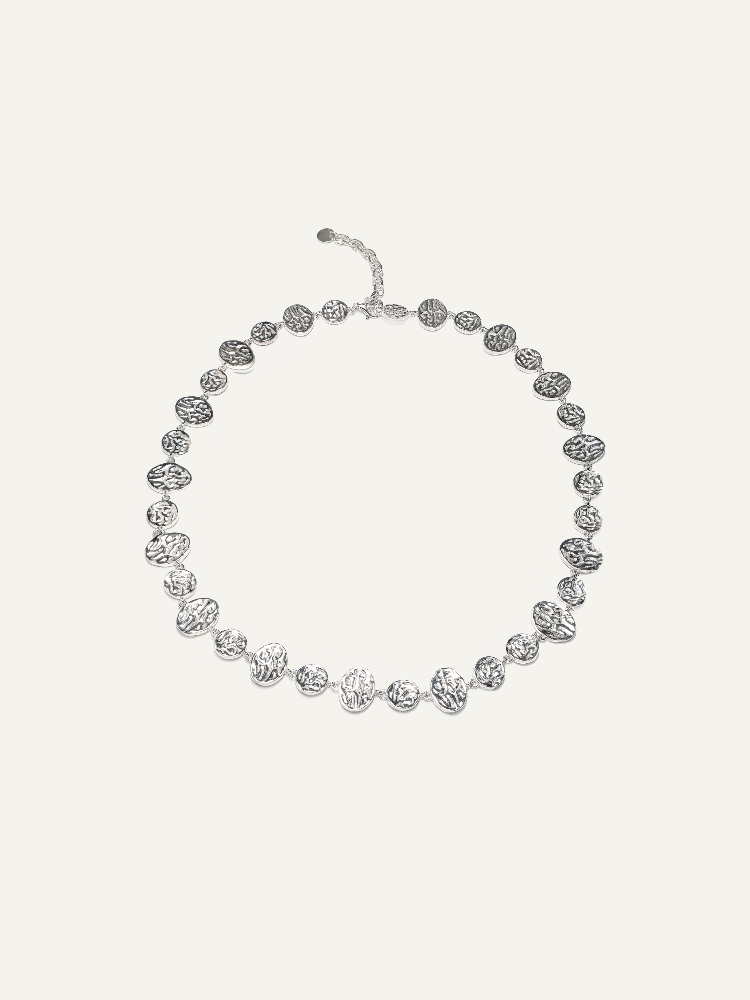 A silver-toned beaded necklace with textured, organic-shaped beads and a matching extender clasp, displayed on a light cream background.