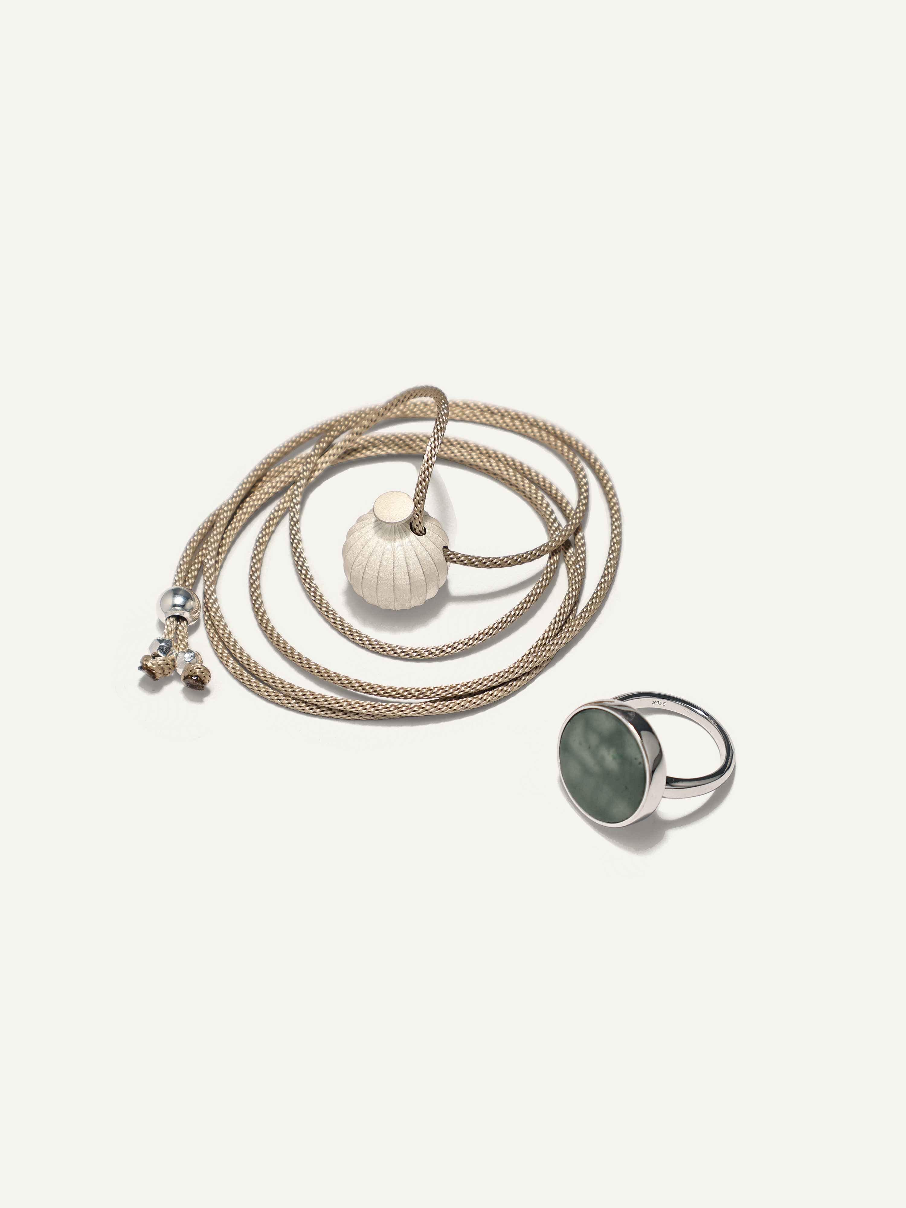 A bohemian-style jewelry set featuring a thick beige braided rope necklace with a white vase pendant and small silver beads, alongside a silver signet ring with a round green jade, displayed on a light cream background.